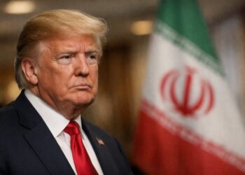 Donald Trump en primer plano con bandera de Irán desenfocada detrás.