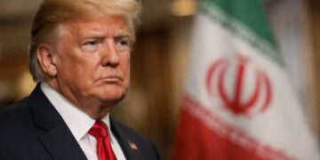 Donald Trump en primer plano con bandera de Irán desenfocada detrás.
