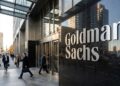 Entrada de la sede de Goldman Sachs con el letrero 'Goldman Sachs' en letras plateadas. Hombres de negocios con trajes caminan por la acera.