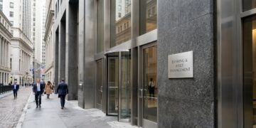 Entrada de edificio moderno con puerta giratoria y placa de 'Banking & Asset Management'. Peatones caminan por la calle mojada.