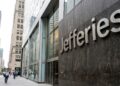 Entrada de la oficina de Jefferies en Nueva York con el nombre en letras grandes de metal sobre piedra. Peatones caminan por la acera.