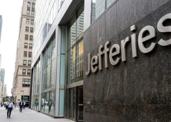 Entrada de la oficina de Jefferies en Nueva York con el nombre en letras grandes de metal sobre piedra. Peatones caminan por la acera.