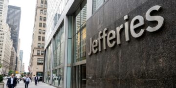 Entrada de la oficina de Jefferies en Nueva York con el nombre en letras grandes de metal sobre piedra. Peatones caminan por la acera.