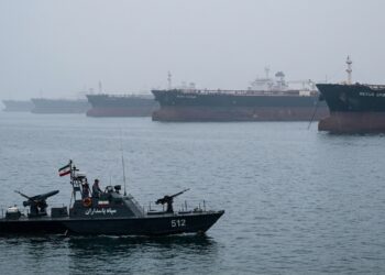 Lancha militar de Irán con bandera navega cerca de grandes buques petroleros en el mar brumoso de Hormuz.