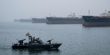 Lancha militar de Irán con bandera navega cerca de grandes buques petroleros en el mar brumoso de Hormuz.