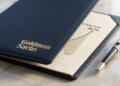 Carpeta azul de Goldman Sachs con logo dorado, abierta mostrando gráfico de 26.2% y pluma.