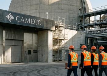 Trabajadores con chalecos naranjas y cascos caminan frente a la entrada de Cameco con una gran estructura de concreto al fondo.