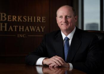 Greg Abel en sala corporativa junto al logo de Berkshire Hathaway