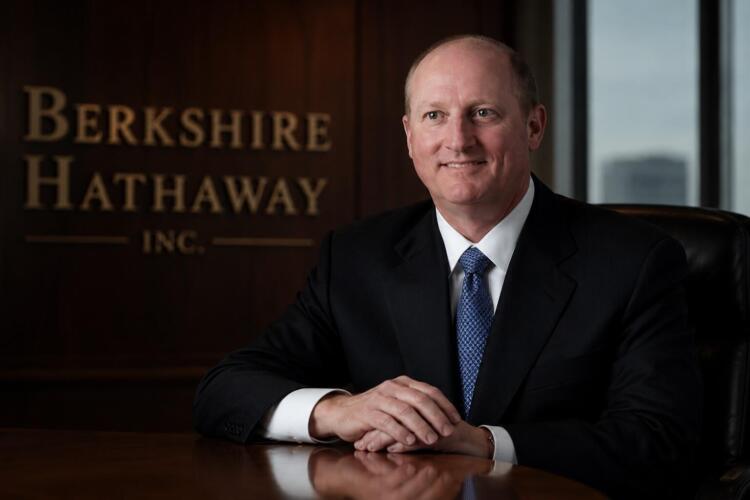 Greg Abel en sala corporativa junto al logo de Berkshire Hathaway