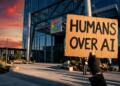 Mano enguantada con cartel "HUMANS OVER AI" frente a edificio de Google al atardecer.