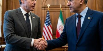 Antony Blinken y Hossein Amir-Abdollahian dándose la mano con banderas de EE.UU. e Irán detrás.