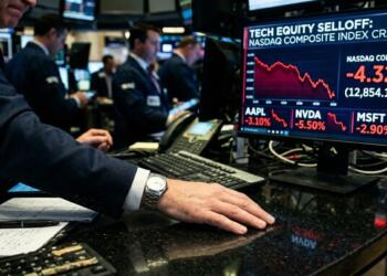 Primer plano de brazo de broker con reloj sobre escritorio, observando pantalla de crash del Nasdaq con gráficos en rojo.