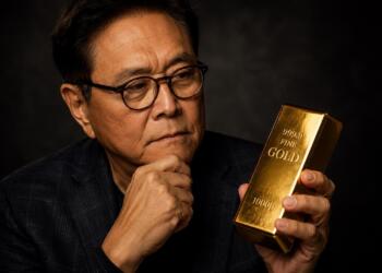 Robert Kiyosaki observa una barra de oro con expresión reflexiva.
