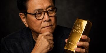 Robert Kiyosaki observa una barra de oro con expresión reflexiva.