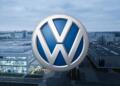 Logo de Volkswagen frente a una planta industrial parcialmente en penumbra.