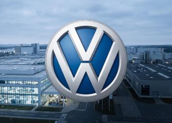 Logo de Volkswagen frente a una planta industrial parcialmente en penumbra.