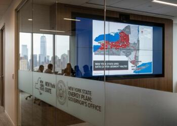 Oficina con una pantalla que muestra un mapa de la red eléctrica de Nueva York, indicando la suspensión del segmento de centros de datos, vista a través de una pared de cristal con logotipos del estado.
