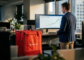 Hombre de pie frente a una pantalla con gráficos de DoorDash y una bolsa térmica roja.