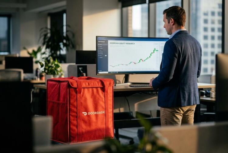 Hombre de pie frente a una pantalla con gráficos de DoorDash y una bolsa térmica roja.