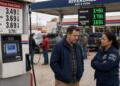 Un hombre y una trabajadora conversan en una gasolinera con precios de combustible visibles.