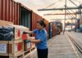 Mujer inspecciona cajas de madera con etiquetas de drones en una terminal portuaria.