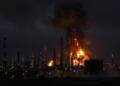 Vista nocturna de una refinería de petróleo con grandes llamas y una columna de humo.