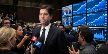 Kevin Warsh rodeado de reporteros que le extienden micrófonos frente a pantallas.