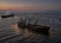 Barcos cargueros humeantes y dañados navegando en el mar durante un atardecer nublado.