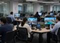Varios trabajadores en una oficina moderna observan computadoras y teléfonos móviles.