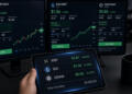 Una persona sostiene una tableta con precios de criptomonedas frente a varias pantallas.
